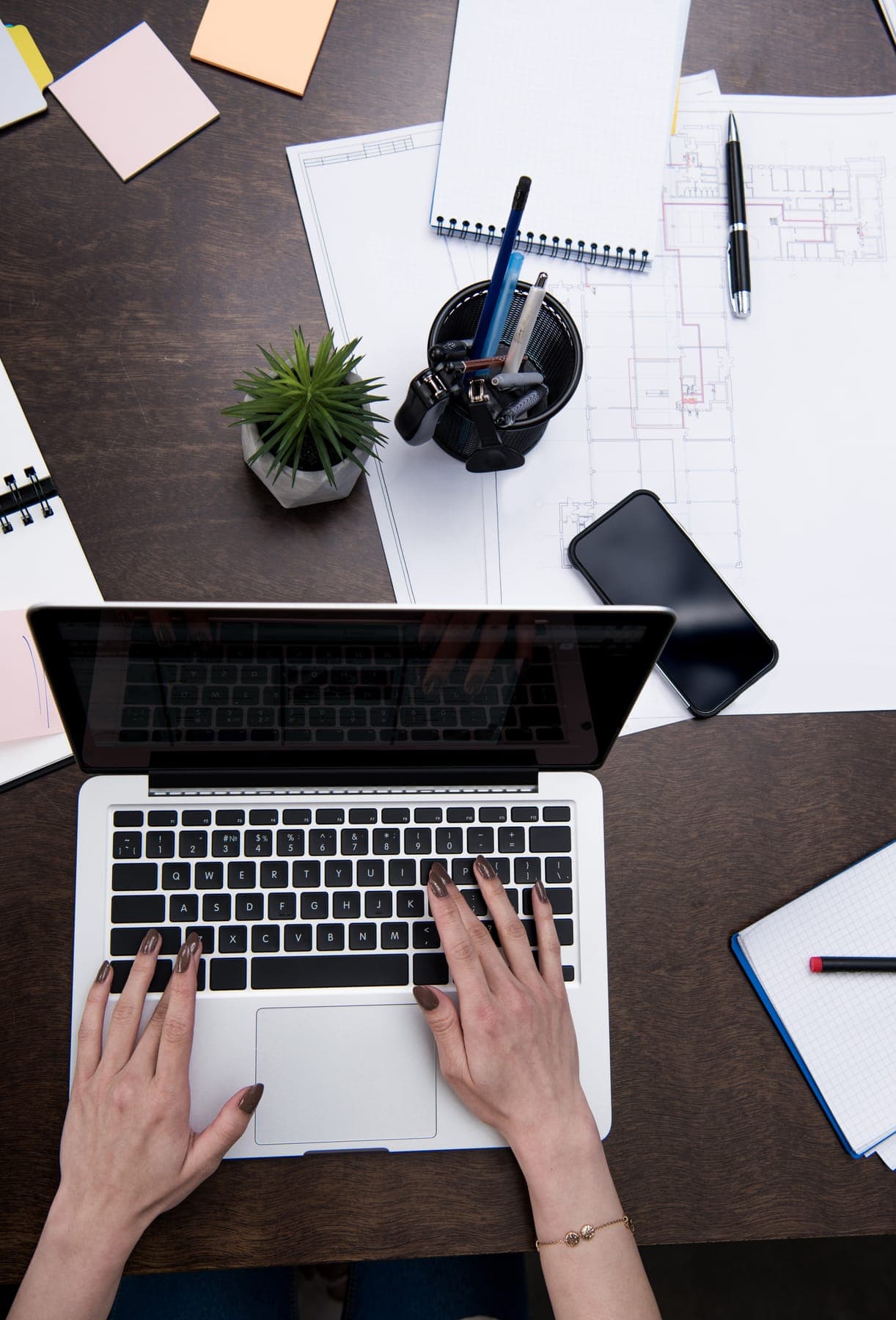 Hands typing on a laptop at a desk covered with plans and notes